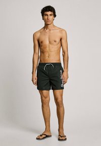 Juodi maudymosi šortai su baltais logotipo akcentais, elastinga liemens juosta su raišteliu, pagaminti iš lengvos medžiagos. Dėvimi su juodomis šlepetėmis.