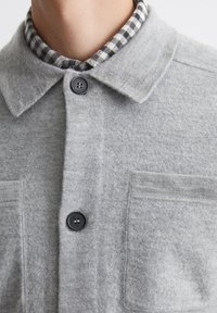 Gray wool shirt featuring a pointed collar, black buttons, and a black and white checkered inner lining with a single chest pocket.