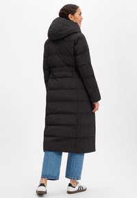 Zwarte pufferjas met capuchon, taille met riem en doorgestikte textuur. Draagt deze over lichtblauwe jeans met wijde pijpen en witte sneakers met zwarte strepen.