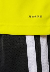 Koszulka sportowa AEROREADY ma jasną żółtą górę, oddychające czarne szorty z białymi bocznymi paskami oraz gładkie tekstury materiału.