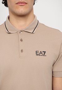 Beige polo shirt with a textured fabric, black trim on the collar, three buttons, and a black EA7 Emporio Armani logo on the chest.