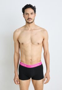 Boxer-briefs noirs avec une ceinture rose arborant le logo "Lacoste". Le tissu semble doux, avec une coupe ajustée et sans motifs ou accents visibles.