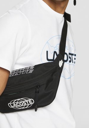 Bolso de hombro de tela negra con un compartimento con cremallera, con texto blanco "LACOSTE" y detalles de logo. Llevado sobre una camiseta gráfica blanca.