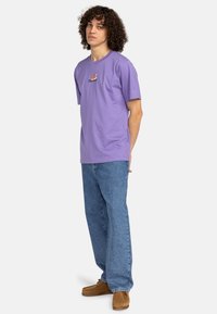 Camiseta de algodón morada de manga corta y cuello redondo, con un pequeño logotipo en el pecho. Combinada con unos pantalones vaqueros de mezclilla azul claro.