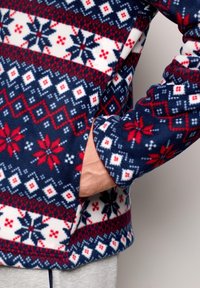 Mano parzialmente in tasca di una giacca in pile con motivi nordici a fiocchi di neve e diamanti in rosso, bianco e blu.