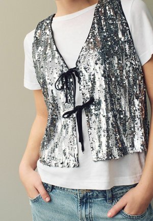 Osoba ubrana w białą koszulkę i niebieskie dżinsy, z srebrną kamizelką z cekinów zawiązaną czarnymi wstążkami na kokardki na koszulce.