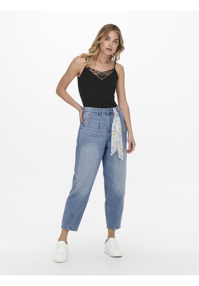 Top en dentelle noire avec bordure, associé à un jean en denim bleu clair taille haute, avec une écharpe à motifs floraux nouée à la taille et des baskets blanches.