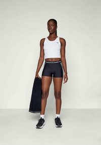 Fehér sportos crop top, fekete kompressziós rövidnadrág "Under Armour" derékkal, fekete törölközőt tart, fekete sportcipőt visel. Semleges háttér.
