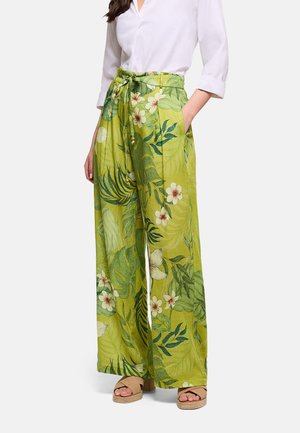 Mujer con pantalones anchos verdes con estampado floral y bolsillos y una camisa blanca abotonada, combinados con sandalias beige de cuña.