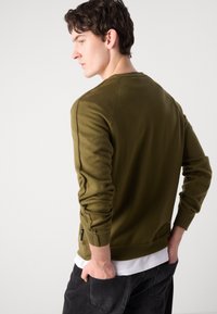 Olijfgroene trui met een gestructureerde breiwijze, ronde halslijn, geribbelde manchetten en onderzoom. Zwarte label aan de zijnaad, gedragen over een witte shirt.