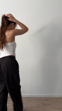 Hosszú hajú nő, aki hátraköti a haját, fehér pánt nélküli felsőt és fekete nadrágot visel, egy fehér, textúrált falnak és fából készült padlón áll.