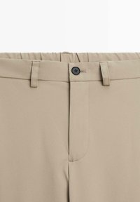 Pantalon beige avec une taille à passants de ceinture, bouton noir, et panneau en stretch élastique à l'arrière.