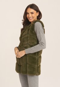 Olive green faux fur vest with quilted design, featuring plush texture and a relaxed fit, worn over a gray long-sleeve top.
