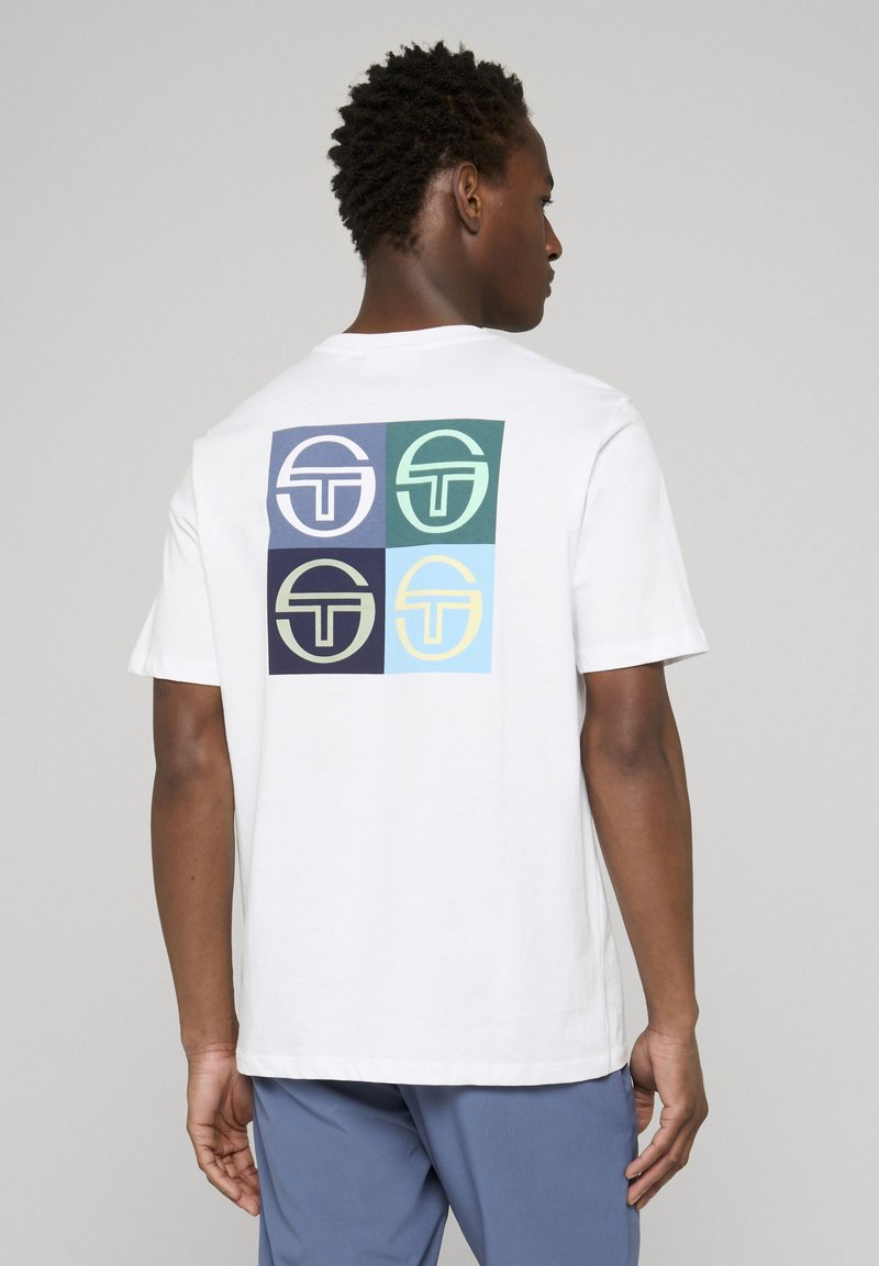 Fehér pólót viselő férfi, aki négy színes négyzet alakú logót visel a hátán, valamint kék nadrágot, egyszerű háttér előtt, háttal a nézőnek.