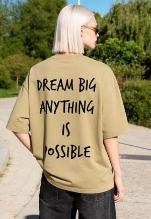 Persoon met kort licht haar die een zonnebril en een beige shirt met de tekst "DREAM BIG ANYTHING IS POSSIBLE" draagt, staand buiten op een zonnige dag.