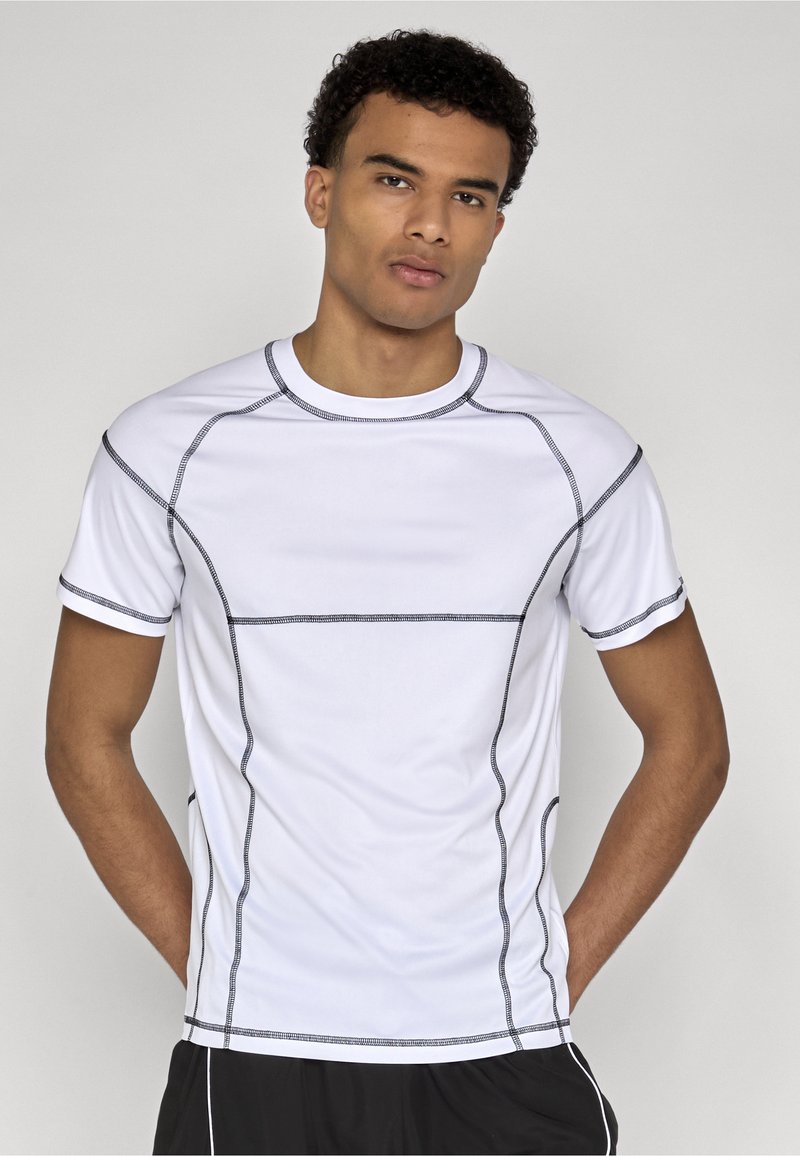Jeune homme aux cheveux courts et bouclés portant un t-shirt de sport blanc avec des coutures noires, debout les mains derrière le dos.