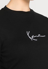Schwarzes geripptes T-Shirt mit einem weißen gestickten Signatur-Logo auf der linken Brustseite, das einen runden Ausschnitt und einen figurnahen Schnitt zeigt.
