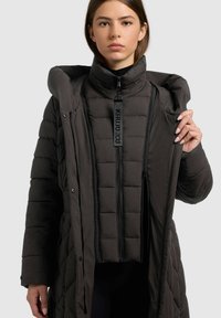 Fiatal nő fekete, rétegezett puffer dzsekit visel, az outer réteget nyitva tartja, hogy megmutassa a belső, cipzárral záródó réteget, amely márkás húzótárral rendelkezik.