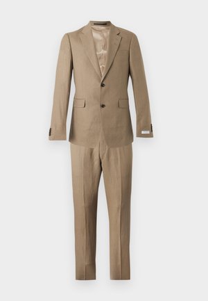 Mladý muž stojaci s rukami v kapsách, oblečený v béžovom obleku, modrozeleniacej košeli a pieskovo hnedej semišovej obuvi so šnúrkami, na jednoduchom pozadí.