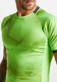 Camiseta atlética verde con un patrón geométrico texturizado, de mangas cortas y un cuello negro. Presenta un pequeño logotipo blanco en el pecho.