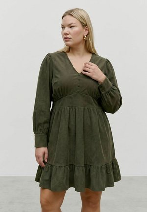 Femme aux cheveux blonds clairs portant une robe longue manches longues de couleur olive avec des boutons et un ourlet à volants, debout devant un fond clair uni.
