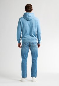 Sudadera con capucha de color azul claro con un acabado texturizado, combinada con jeans azules claros y zapatillas blancas, vista desde la parte posterior.