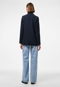 Personne aux cheveux courts vue de dos, portant un blazer bleu marine à fines rayures, un jean large bleu clair et des chaussures noires sur un fond blanc.