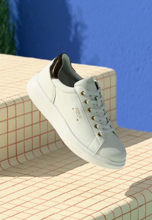 Sneaker bianco con dettagli in cuoio marrone e occhielli dorati, appoggiato su gradini in piastrelle beige con uno sfondo di muro blu.