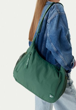 Persona con chaqueta de mezclilla que lleva un bolso verde grande de Reebok con cremallera y bolsillos laterales de malla.