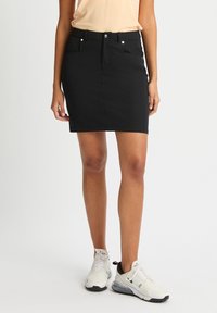 Mini gonna in denim nera con una forma aderente, passanti per cintura, tasche frontali e dettagli in metallo argento. Indossata con sneakers bianche.