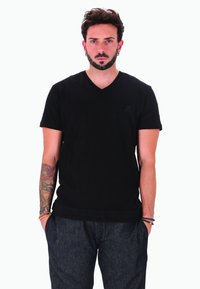 Uomo con capelli ricci scuri e barba, in piedi di fronte, indossa una maglietta nera a scollo a v, pantaloni scuri, braccialetti e un tatuaggio floreale sul braccio sinistro.