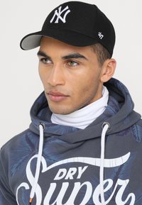 Casquette de baseball noire avec logo New York blanc, portée avec un sweat à capuche gris arborant un motif de feuilles de palmier bleues et des cordons blancs.