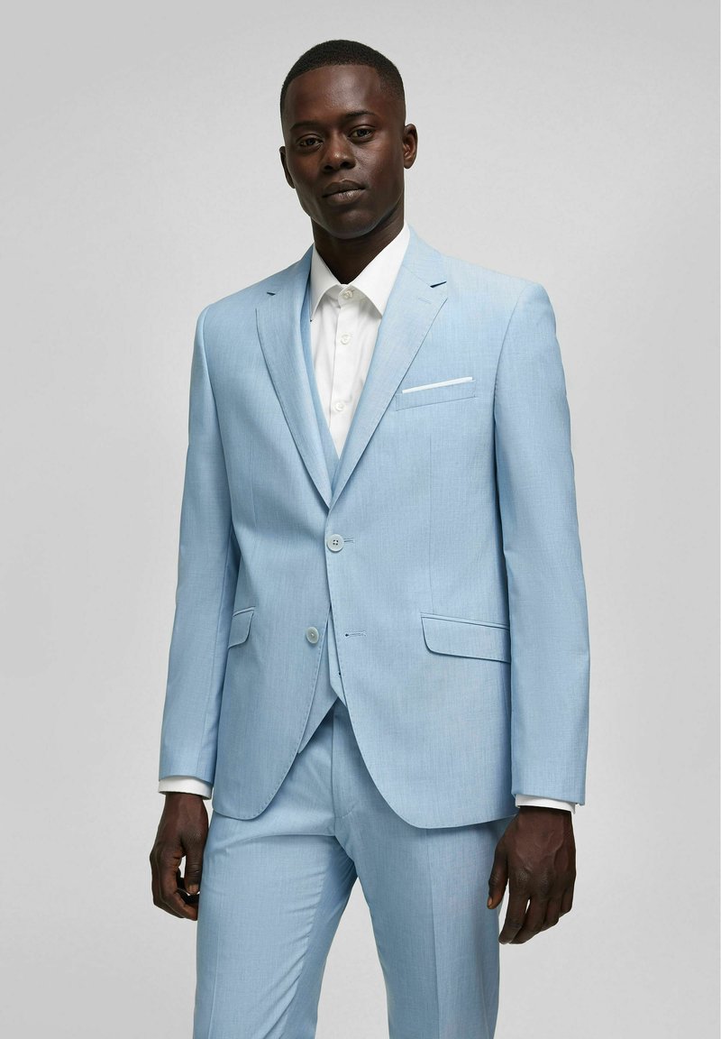 Helles blaues Sakko mit tailliertem Schnitt, strukturiertem Stoff, Einknopf-Verschluss und einem weißen Hemd darunter. Auffälliges Taschendetail an der Jacke.