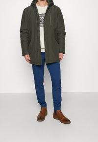 Veste imperméable verte avec une poche poitrine zippée, portée par-dessus un pull crème à motif géométrique, associée à un pantalon bleu et des bottes marron.