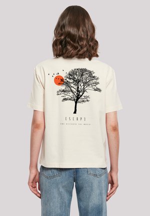 Woman with shoulder-length brown hair wearing cream T-shirt with black tree, orange sun, flying birds, and text "ESCAPE and discover the world" on back.
