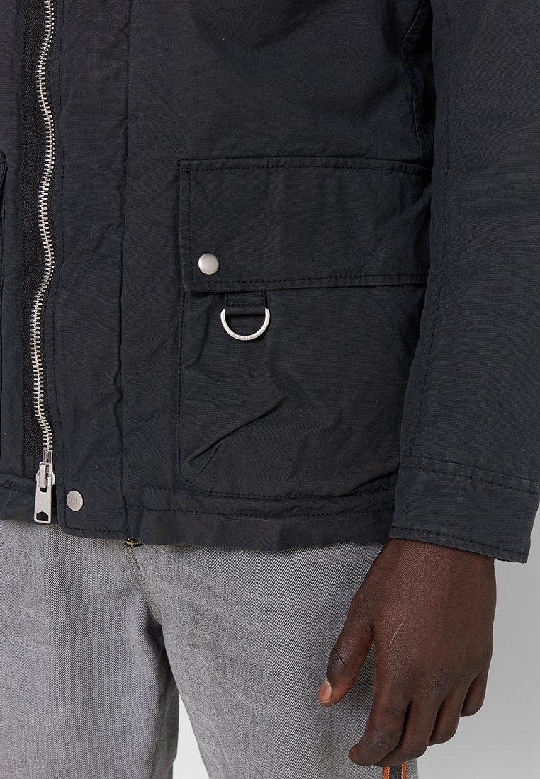 Schwarze Utility-Jacke mit Reißverschluss, Fronttasche und Ringakzent. Der Stoff ist strukturiert und hat ein mattes Finish.