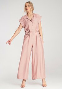 Jasnoróżowy kombinezon z miękkiego materiału, z krótkimi rękawami, kołnierzykiem, guzikami i ozdobnym wiązanym detalem w postaci kwiatu w talii.