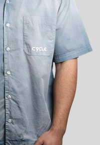 Camicia di cotone azzurro chiaro a maniche corte realizzata in tessuto tessuto. Presenta una tasca sul petto e il logo "CYCLE" in bianco sulla tasca. Design con bottoni davanti.