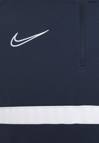 Granatowa bluza z zamkiem do połowy, wykonana z gładkiego materiału, z białym poziomym paskiem i małym białym logo Nike na lewej piersi.