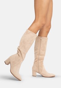 Botas de ante beige hasta la rodilla con tacón bloque, que presentan una silueta curvada y una cremallera lateral para un fácil uso. Textura suave y diseño ajustado.