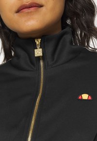 Gros plan d'une personne portant une veste noire zippée avec une fermeture éclair dorée et un petit logo en demi-cercle rouge et orange sur la poitrine.
