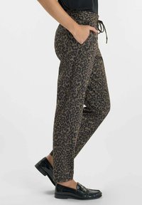 Vrouw in bruine en zwarte luipaardprint broek met elastische tailleband en zwarte instappers, staande met één hand in haar zak.