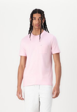 Lyserød bomulds T-shirt med korte ærmer, rund halsudskæring og en brystlomme. Model iført lys grå sweats, stående foran en hvid baggrund.