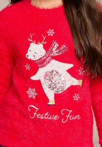 Röd fluffig tröja med en paljetterad isbjörn som bär en halsduk, snöflingor och texten "Festligt Kul".