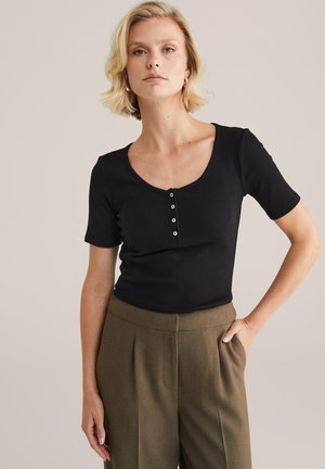 Top noir à manches courtes avec un col en U et une patte de boutonnage à cinq boutons, fabriqué en tissu côtelé. Associé à un pantalon marron ajusté.