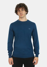 Maglione blu a maniche lunghe realizzato in tessuto morbido, con scollo rotondo e un piccolo logo sul lato sinistro del petto. Texture liscia, design aderente.
