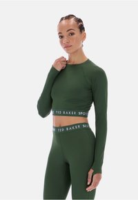 Žalias trumpas viršelis su ilgomis rankovėmis, pasižymintis prigludusia išvaizda ir logotipo juosta apatiniame krašte, derinamas su atitinkančiomis aukštu juosmeniu kelnėmis.