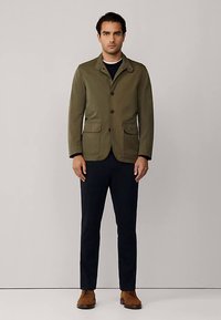 Maslinasto zelena jakna s ovratnikom, zatvaranjem na prednje gumbe, dva džepa na prsima i opuštenim krojem. Uparena s tamnim hlačama i smeđim cipelama.