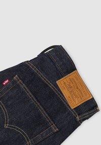 Coin de jeans Levi's en denim bleu foncé avec poche arrière visible, couture orange, étiquette rouge Levi's et étiquette de marque en cuir marron à la ceinture.