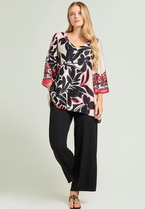 Kvinde iført beige tunika med sorte og røde blomsterprint, sorte bukser med vide ben og sorte pyntede sandaler, stående mod ensfarvet baggrund.